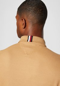 Polo beige en tissu texturé, avec un col accentué par des rayures rouges, blanches et bleues au niveau de l'encolure arrière.