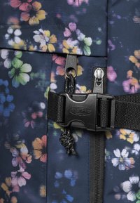 Floral-patterned backpack in dark fabric features black hardware, zippers, and an adjustable strap with a plastic buckle and cord pull.