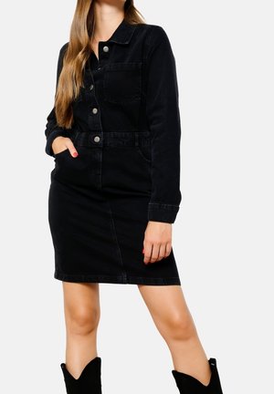 Frau trägt ein schwarzes Denim-Kleid mit Knöpfen, langen Ärmeln und Taschen, kombiniert mit schwarzen Stiefeletten, vor weißem Hintergrund.