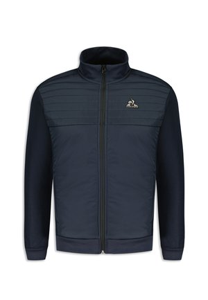 Veste zippée marine avec des panneaux rembourrés sur la poitrine, col et poignets côtelés. Présente un logo sur la partie gauche de la poitrine. Texture lisse.