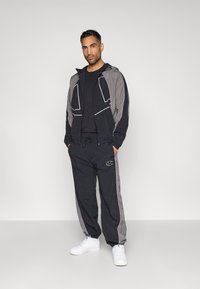 Chándal en negro y gris con chaqueta con cremallera y capucha, y pantalones con cintura elástica. Acento de logotipo blanco en los pantalones. Zapatillas blancas.