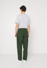 Uomo in maglietta grigia chiara, pantaloni verde scuro con tasche posteriori, e sneakers bianche in piedi di fronte a un muro bianco semplice.