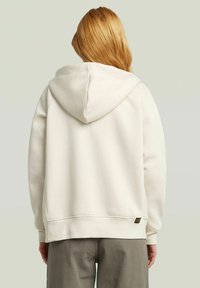 Person with long red hair wearing a plain light beige hoodie and gray pants, viewed from behind against a light gray background.