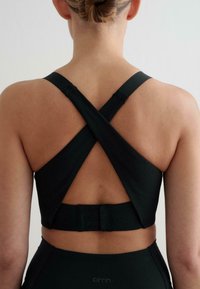 Černá sportovní podprsenka s ribbed texturou, s překříženými ramínky a háčkovým zapínáním na zadní straně. Hladká tkanina a strukturovaný design.