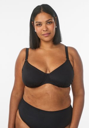 Itsevarma nainen, jolla on keskiruskea iho ja suorat mustat hiukset, pukeutuneena mustaan rintaliiviin ja siihen sopiviin mustiin alushousuihin, seisoo valkoista taustaa vasten.