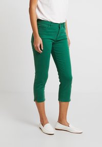 Pantalon capri vert en coton, avec une texture douce, une coupe slim, des fermetures éclair aux chevilles et une taille mi-haute. Assorti à des chaussures blanches faciles à enfiler.