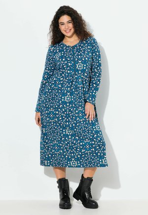 Langärmliges, knielanges Kleid in Blau mit floralem Muster, das weiße und türkise Akzente aufweist. Kombiniert mit schwarzen Kampfstiefeln.