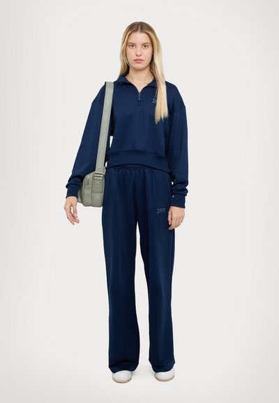 Navy pullover with a zippered collar and matching pants; both in a relaxed fit, paired with a light green shoulder bag and white shoes.