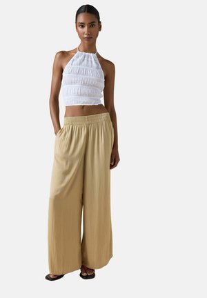 Frau trägt ärmelloses weißes gerafftes Neckholder-Top, lockere helle breite Hose in Hellbraun und schwarze Sandalen vor schlichtem hellem Hintergrund.