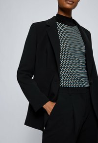 Black tailored blazer featuring a single-button closure, sharp lapels, and a textured patterned shirt in blue and neutral tones underneath.