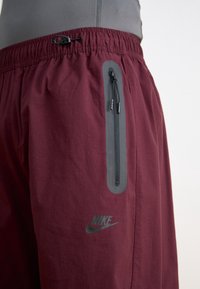 Bordeaux-kleurige sportshorts met een grijze ritszak en een zwart Nike-logo. Voorzien van een elastische tailleband en een gladde stoftextuur.