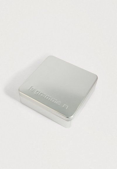 Mazs sudraba metāla trauks ar noapaļotām malām, uz kura vāka ir reljefā uzraksts "le gramme" un kvadrātveida simbols, uz vienkāršas baltas virsmas.