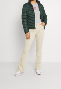 Grön puffjacka med hög krage, ljusgrå t-shirt med röd text och beige utsvängda byxor. Vita sneakers fulländar outfiten.