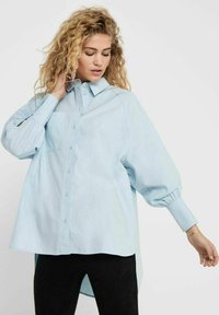 Camicia oversized azzurro chiaro con bottoni, tasca, maniche arrotolate e orlo curvo, realizzata in un tessuto morbido e leggero.