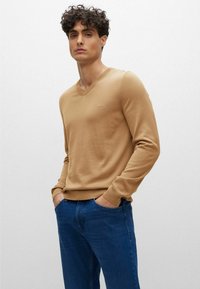 Maglione beige a scollo a V con polsini e orlo a costine, indossato sopra un paio di jeans blu. Nella parte anteriore presenta un sottile dettaglio del logo nella zona del petto.