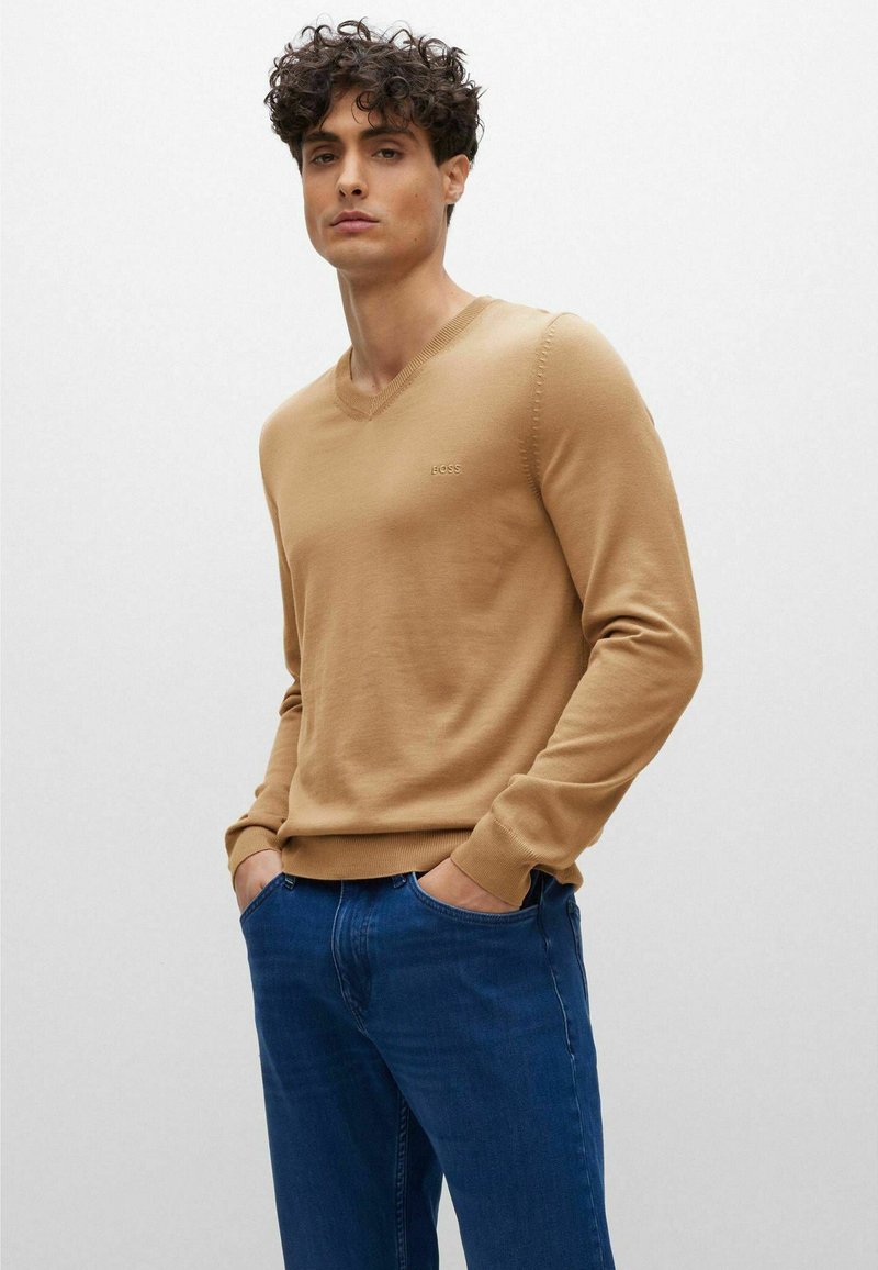 Maglione beige a scollo a V con polsini e orlo a costine, indossato sopra un paio di jeans blu. Nella parte anteriore presenta un sottile dettaglio del logo nella zona del petto.
