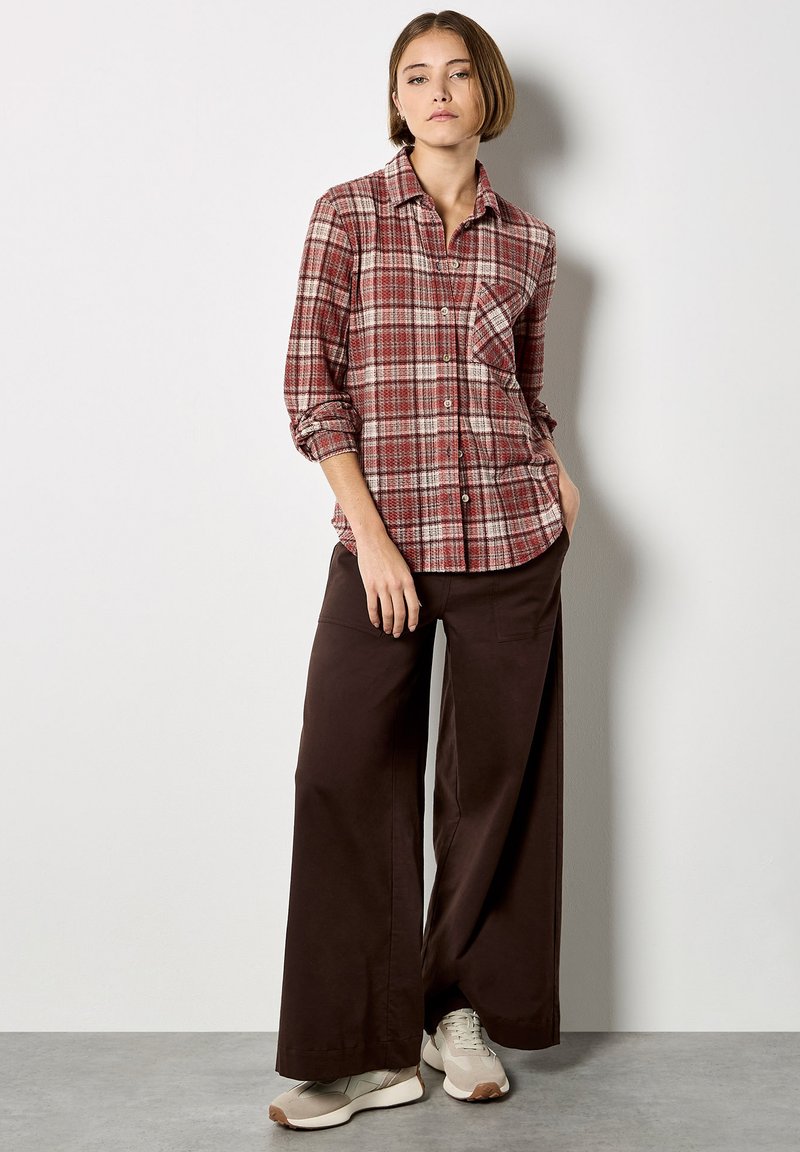 Chemise boutonnière à carreaux rouge et crème avec des manches longues et une poche poitrine, associée à un pantalon large marron et des baskets beiges.