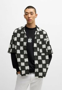 Valkoinen ja musta ruudullinen lyhythihainen paita mustan hupparin päällä, jossa on valkoinen "HUGO"-teksti liekkikoristeilla, yhdistettynä valkoisiin housuihin.