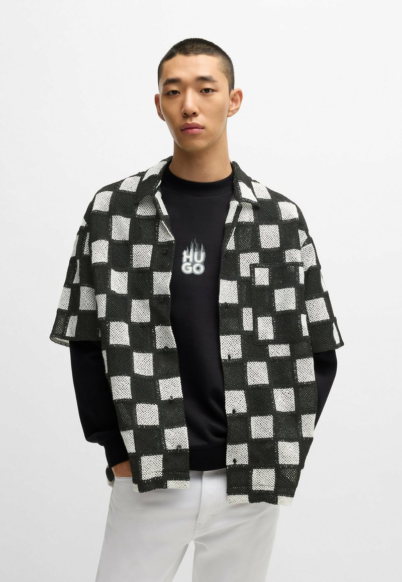 Valkoinen ja musta ruudullinen lyhythihainen paita mustan hupparin päällä, jossa on valkoinen "HUGO"-teksti liekkikoristeilla, yhdistettynä valkoisiin housuihin.