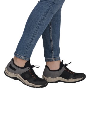 Beine in blauen Jeans, die schwarze und graue Sneaker mit beigen Sohlen tragen, stehen vor einem weißen Hintergrund.