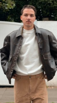 Giubbotto bomber in pelle marrone sopra un maglione bianco, abbinato a pantaloni di velluto a coste color beige. Il giubbotto presenta una chiusura con zip e tasche laterali.