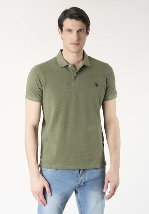 Giovane uomo con capelli scuri che indossa una polo verde oliva e jeans azzurri chiari su uno sfondo bianco uniforme.