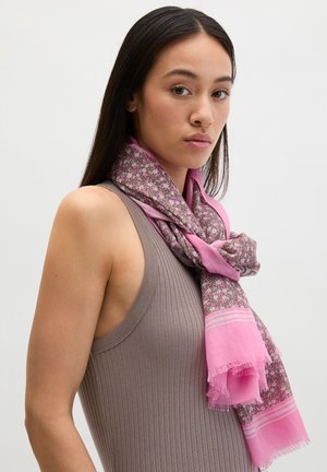 Jonge vrouw met lang donker haar, gekleed in een mouwloos geribd taupekleurig shirt en een roze sjaal met bloemmotief, tegen een effen achtergrond.