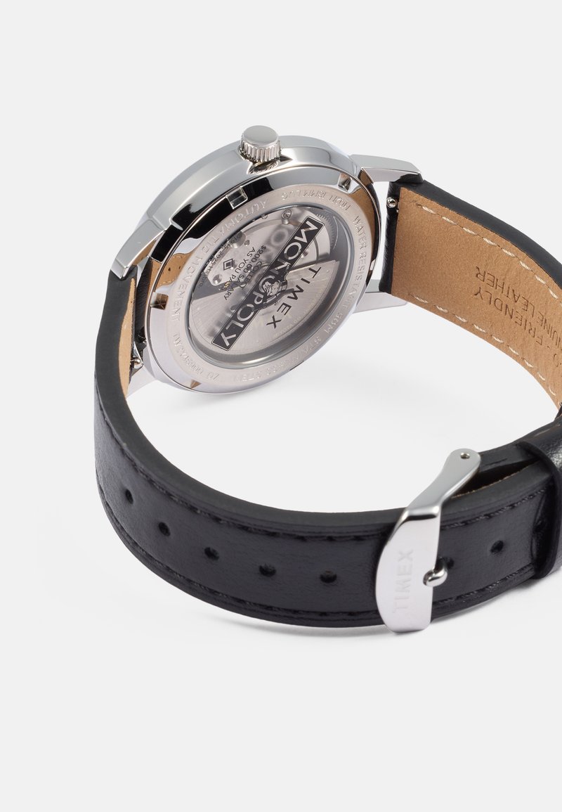 Reloj Timex con caja de acero inoxidable plata, esfera clara y correa de cuero negro. Presenta un cierre de despliegue y una hebilla suave.