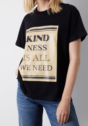 Person trägt schwarzes T-Shirt mit beigem Blocktext "Kindness is all we need" und blaue Jeans, steht vor einfachem Hintergrund.