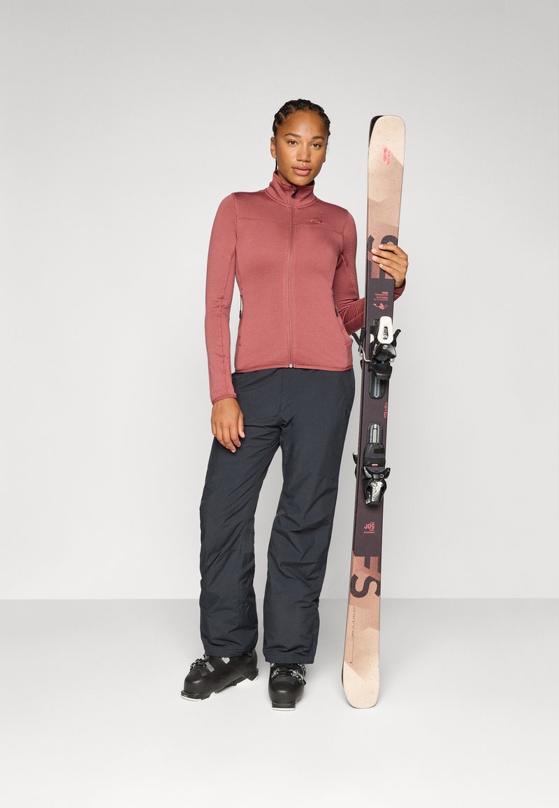 Vrouw in een roodachtige trui en zwarte skibroek houdt ski's vast. Ze staat zelfverzekerd tegen een effen, lichte achtergrond.