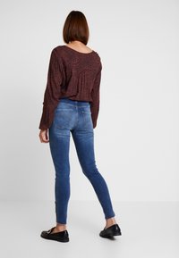 Top de manga larga burdeos con un sutil estampado de leopardo, combinado con jeans ajustados azules que tienen cremalleras en los laterales del dobladillo y zapatos oscuros.