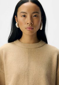 Beige ulltröja med ribbad rund halsringning, med en central stickad söm. Modellen bär stora silverfärgade hoopörhängen.