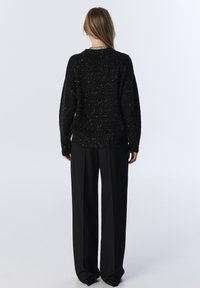 Černý pletený svetr s metalickou nití, volnější střih, žebrované manžety a lem, v kombinaci s širokými černými kalhotami, hladká textura.