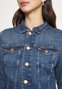 Mörkblå denimjacka med kopparfärgade knapplås, två bröstfickor och klassisk krage. Strukturerad tyg med subtila blekningar.