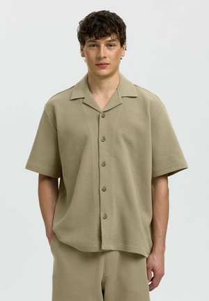 Młody mężczyzna z krótkimi kręconymi włosami nosi beżową teksturowaną koszulę zapinaną na guziki z krótkim rękawem oraz pasujące szorty, stoi na jednolitym tle.