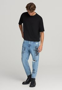 T-shirt nero oversize, jeans strappati azzurri con design in stile graffiti e stivali militari neri. Outfit urbano casual con grafiche distintive.