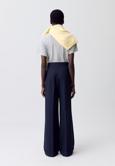 Maglietta grigia a maniche corte, pantaloni larghi a righe navy, e sciarpa gialla chiara poggiata sulle spalle; trame di tessuto lisce visibili.