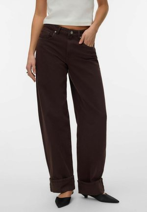 Personne portant un pantalon large taille haute marron foncé avec des revers roulés, un top court blanc et des chaussures noires pointues à talons bas.