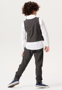 Barn i en gråmönstrad väst över en vit skjorta, i kombination med grå byxor och blå sneakers, sedd bakifrån.