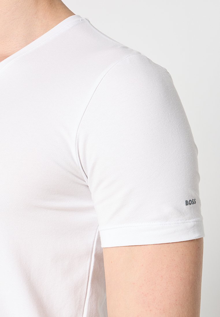 T-shirt blanc en coton à manches courtes avec un design en V. Présente un logo "BOSS" gris sur la manche gauche. Texture lisse, forme ajustée.