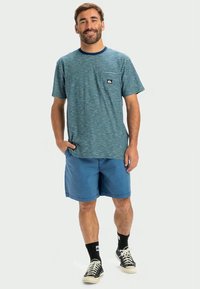 T-shirt azul às riscas com um bolso e logótipo, combinado com calções azuis lisos. Usado com meias pretas e ténis. Ajuste casual.
