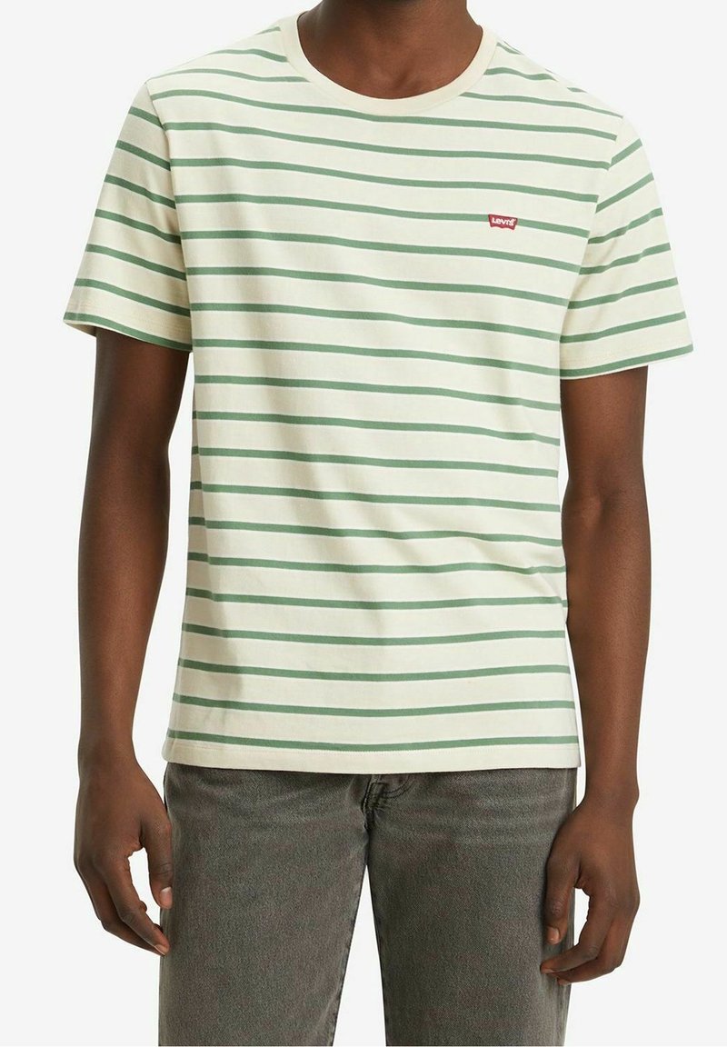 Homme portant un t-shirt à rayures horizontales crème et vert clair avec un petit logo rouge Levi's sur la poitrine et un jean gris.