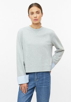 Junge Frau mit braunen Haaren in einem grauen Sweatshirt mit blauen gestreiften Bündchen und blauen Jeans vor einem einfarbigen weißen Hintergrund.