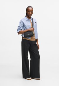 Camicia a scacchi blu e bianca, maglione beige, pantaloni neri larghi, tiene una piccola borsa nera con una tracolla decorata e un portachiavi.