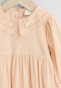 Robe pour bébé de couleur pêche avec un col en dentelle festonné, fermeture à boutons sur le devant et tissu texturé. Présente une jupe plissée et des manches longues.
