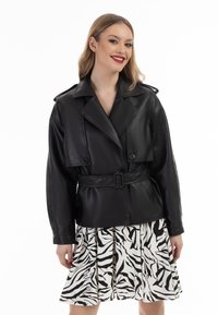 Černá kožená bunda s páskem v pase, přes oversized klopy a knoflíkovými detaily, nošená přes skládací sukni s tygrovaným vzorem.