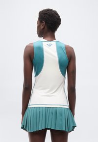 Γυναίκα με κον τά μαλλιά, ντυμένη με αμάνικη λευκή και τιρκουάζ αθλητική μπλούζα και πιετετέ τιρκουάζ φούστα, εμφανιζόμενη από την πίσω πλευρά σε λευκό φόντο.