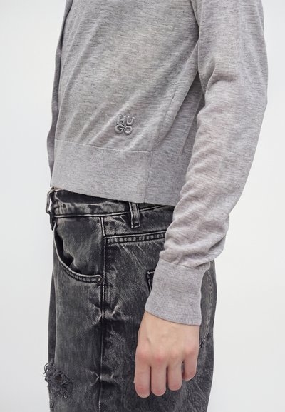 Personne portant une chemise à manches longues gris clair avec le logo "HUGO" et un jean noir déchiré, vue de profil, main détendue le long du corps.