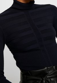 Granatowy sweter z golfem, ozdobiony poziomymi paskami i prążkowanymi detalami przy hemie, noszony z czarnym paskiem w talii.