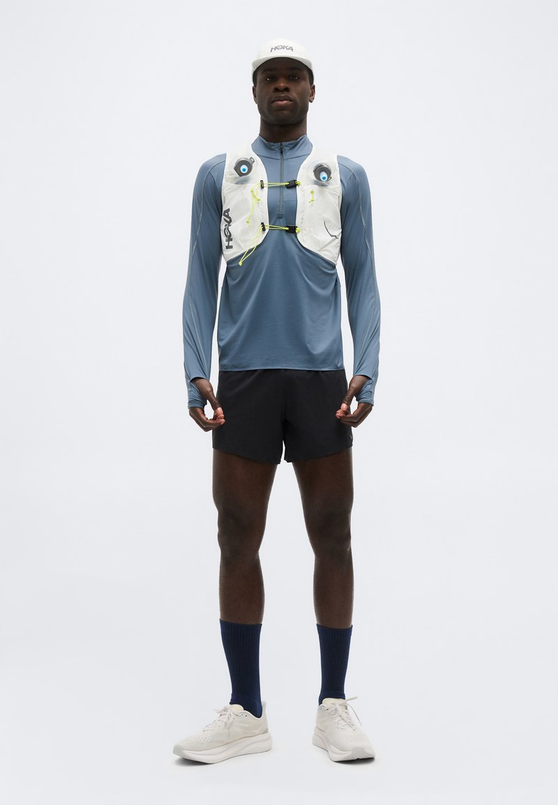 Atleta con gorra blanca, camiseta azul de manga larga, chaleco de hidratación blanco, pantalones cortos negros, calcetines azul marino y zapatillas de running blancas, de pie erguido.